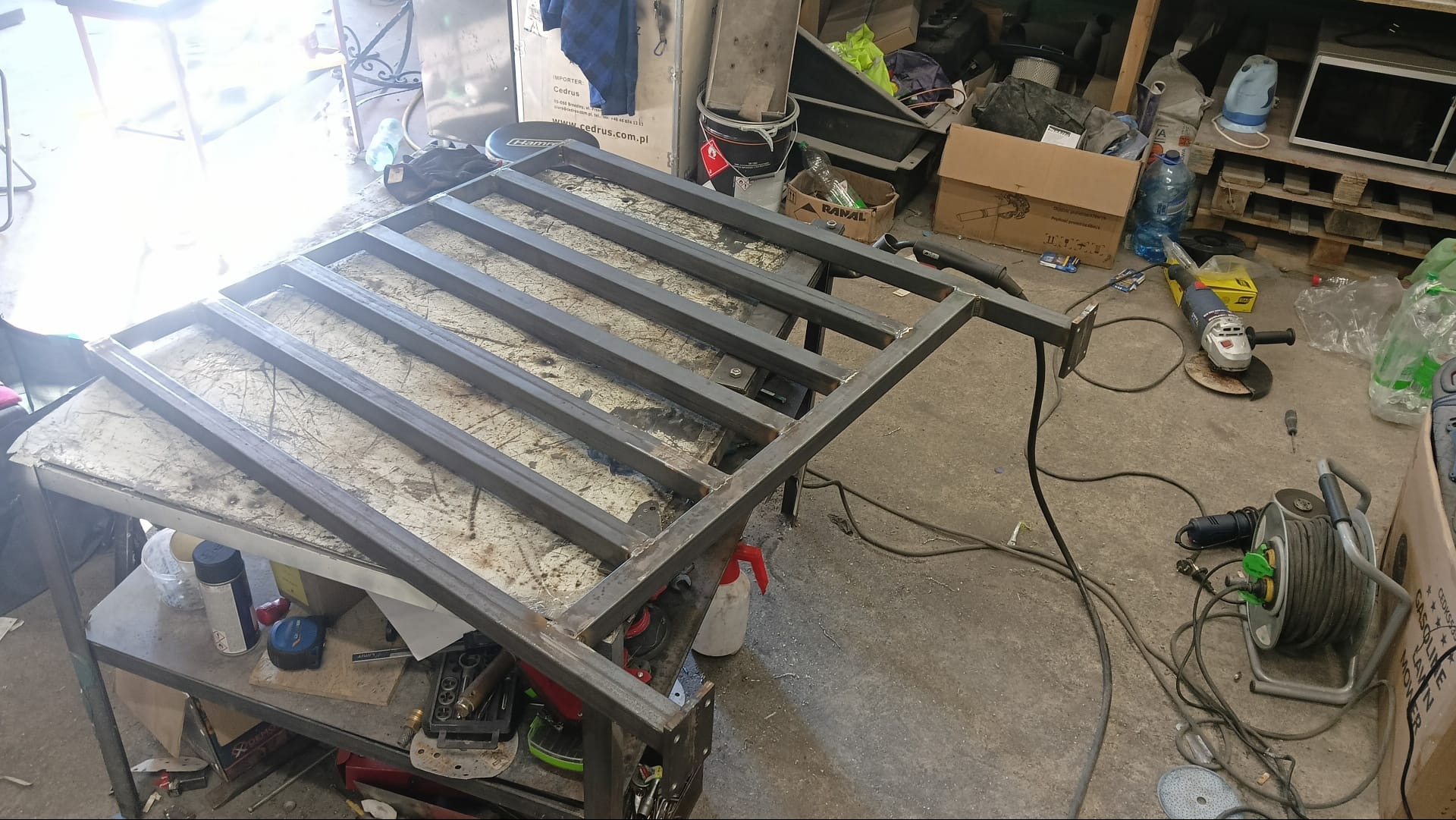 Metalowa konstrukcja z profili kwadratowych, prawdopodobnie rama, na roboczym stole w warsztacie, widoczne ślady spawania, szlifierka kątowa i inne narzędzia w tle.