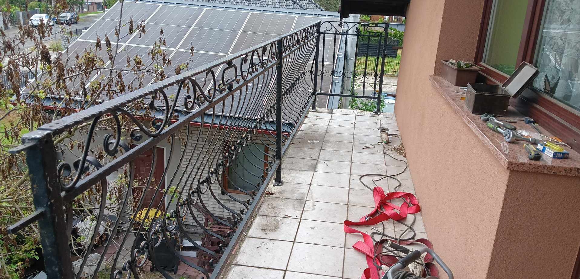 Kuta, czarna balustrada balkonowa z ozdobnymi elementami, widok z góry na balkon w trakcie montażu, widoczne narzędzia i pasy mocujące, w tle panele solarne na dachu budynku.