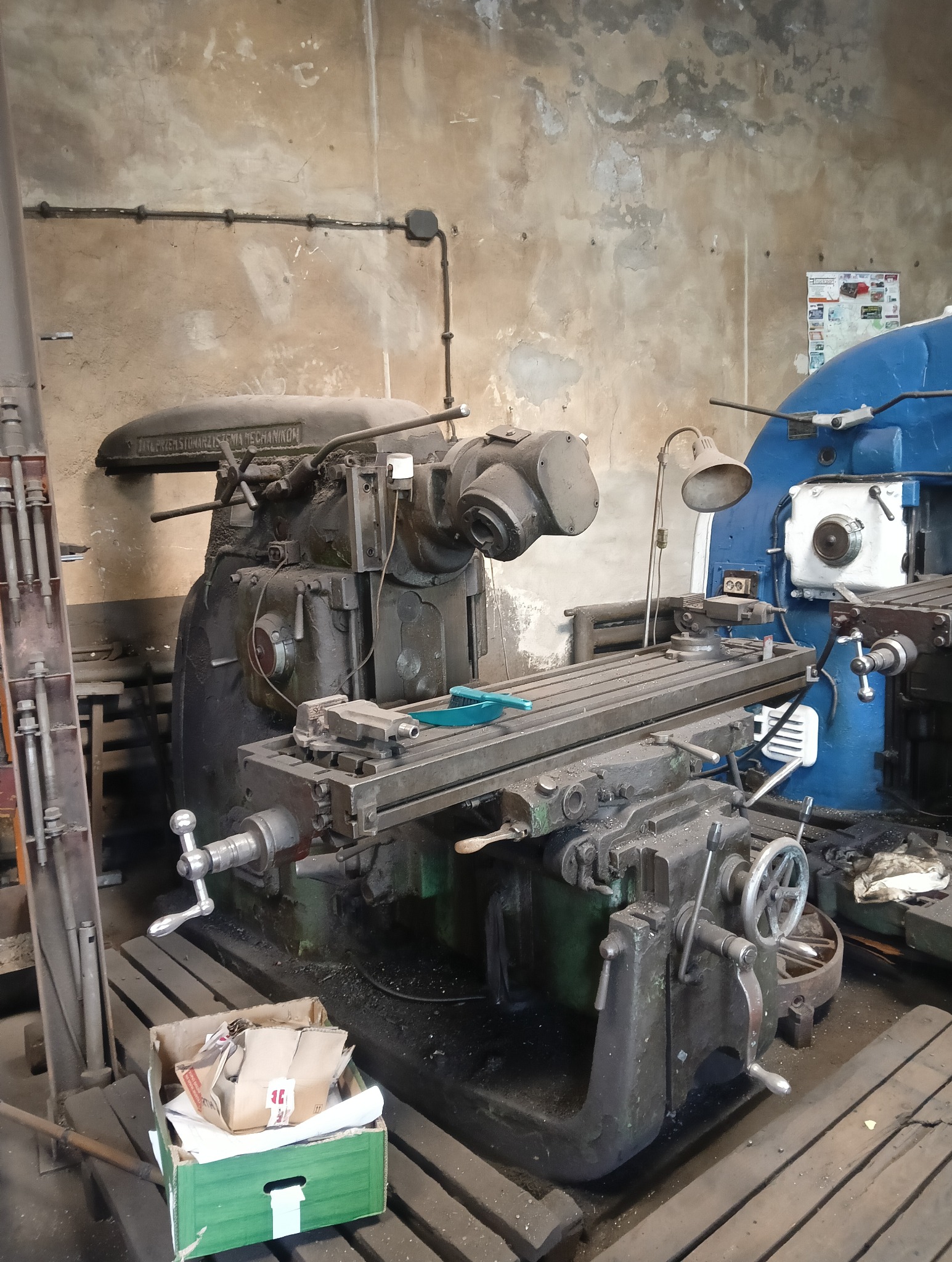 Starodawna frezarka w warsztacie, z widocznym oprzyrządowaniem i skrzynką narzędziową. Na maszynie leży niebieska szczotka do czyszczenia.