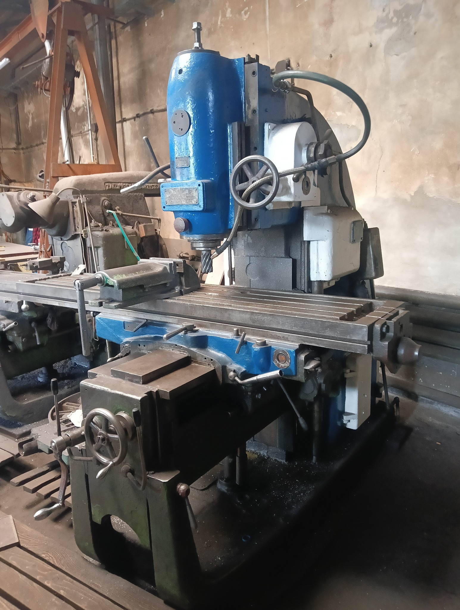 Frezerka pionowa z niebieską głowicą i szarym stołem roboczym w warsztacie. Widoczne pokrętła i uchwyty. W tle inne maszyny i ściany z widocznym zużyciem.