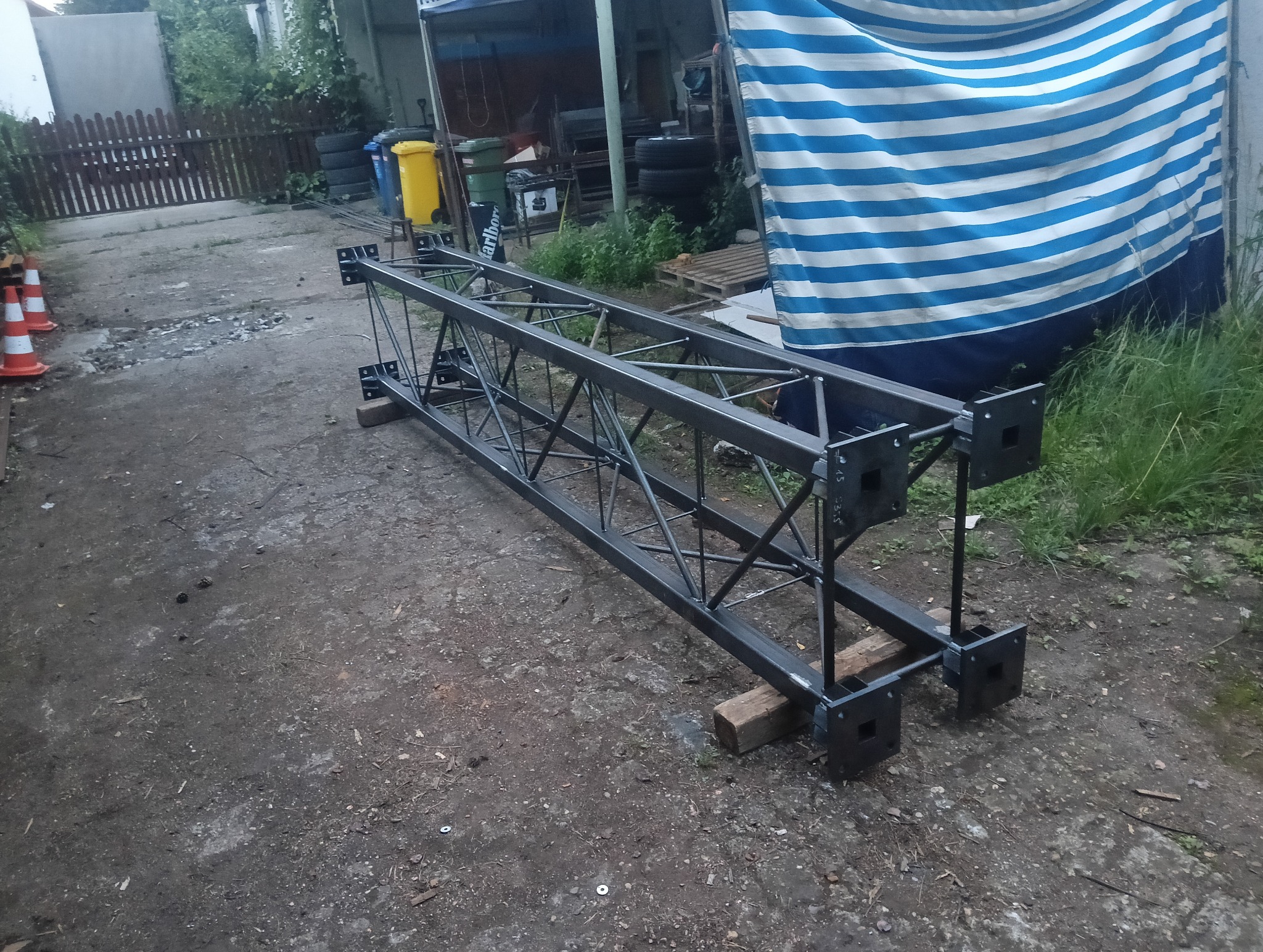 Gotowa, pomalowana konstrukcja metalowa z kratownicą, leżąca na ziemi. Widoczne spawane łączenia i otwory montażowe. W tle budynek i elementy otoczenia.