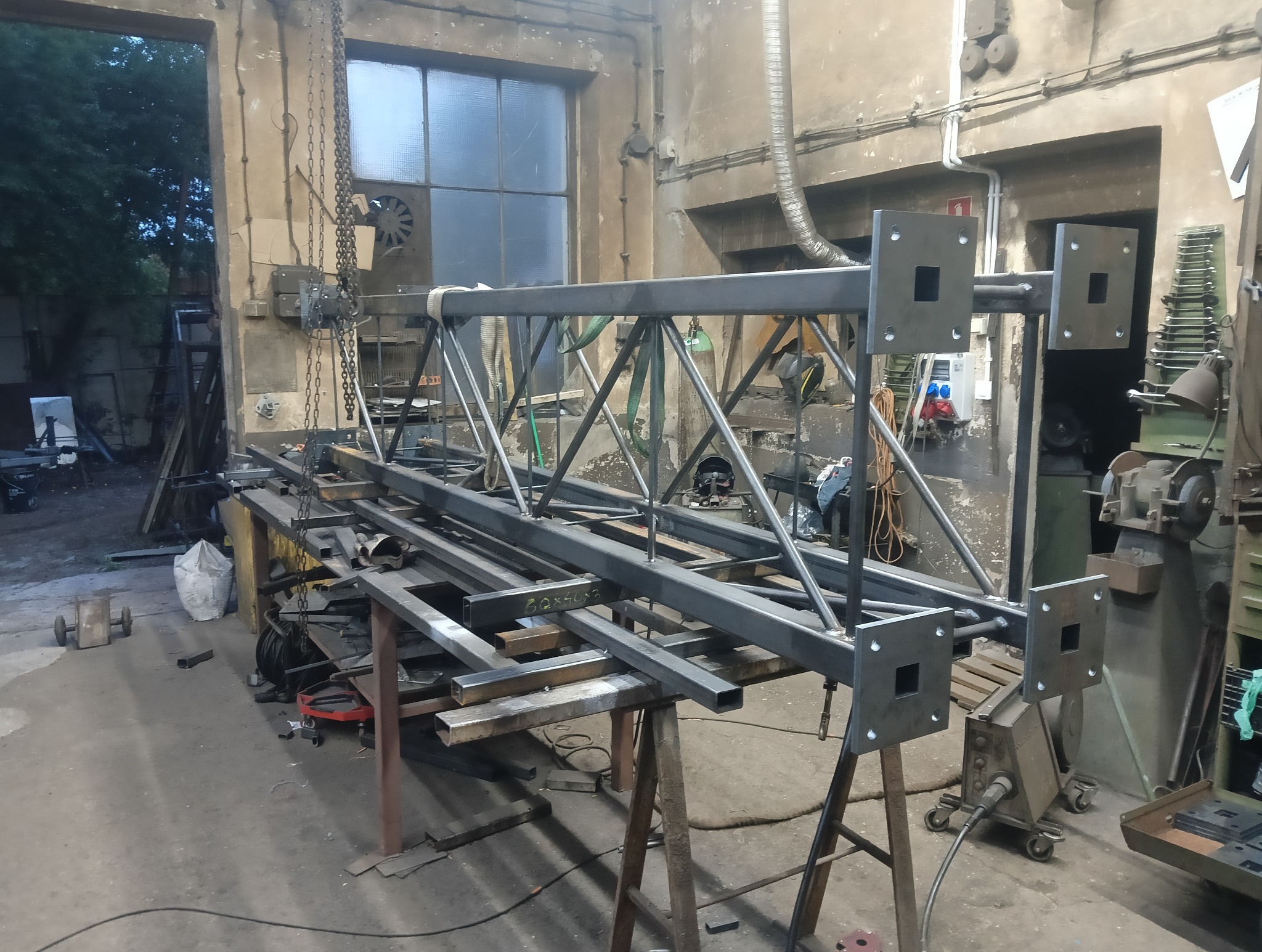 Konstrukcja metalowa z profili i blach w warsztacie, widoczne elementy spawane i przygotowane do montażu. Na stole leżą dodatkowe profile, w tle okno i wyposażenie warsztatu.