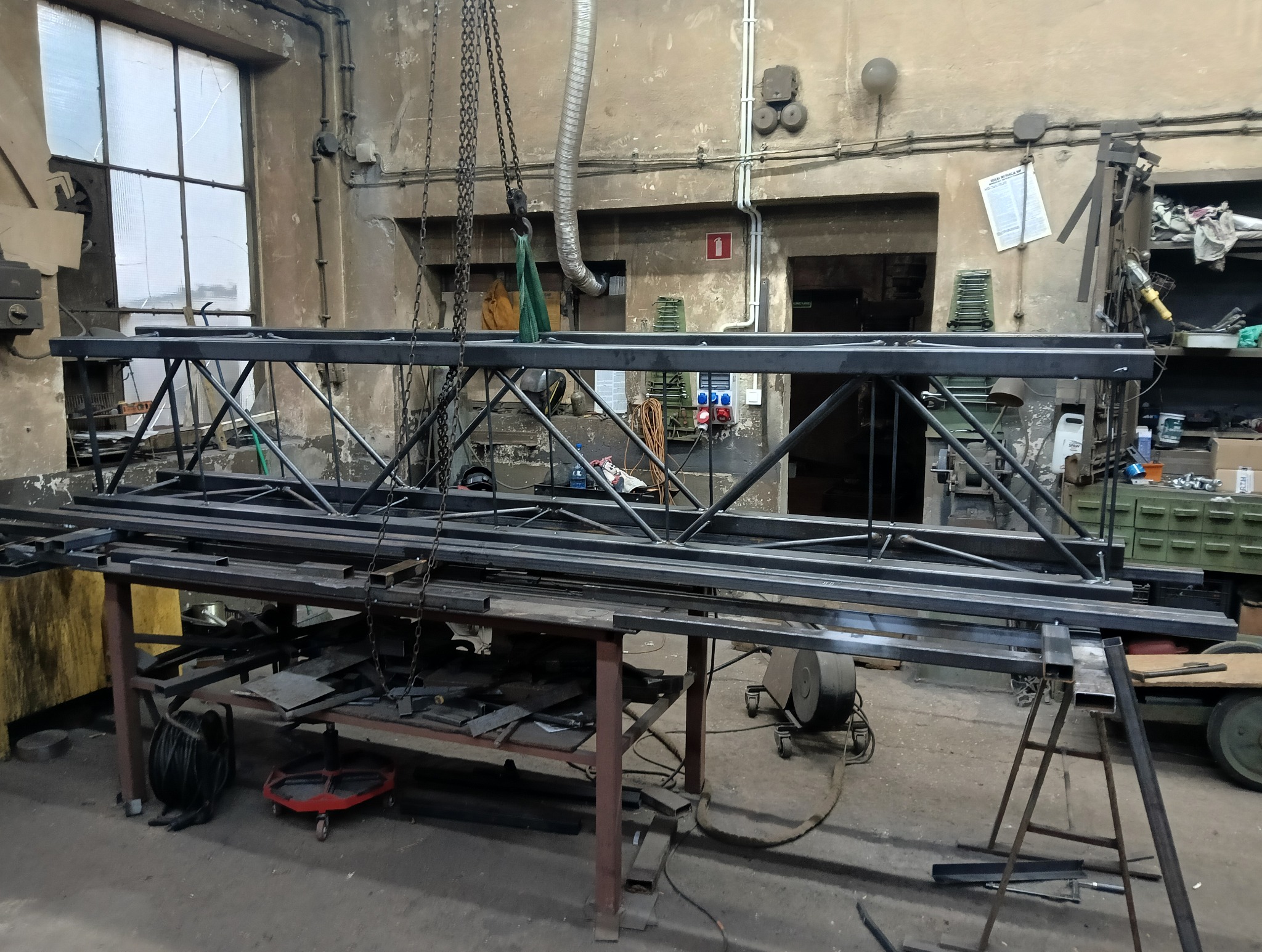 Metalowa konstrukcja kratowa, prawdopodobnie w trakcie spawania, zawieszona nad stołem w warsztacie. Widoczne narzędzia i elementy metalowe w tle.