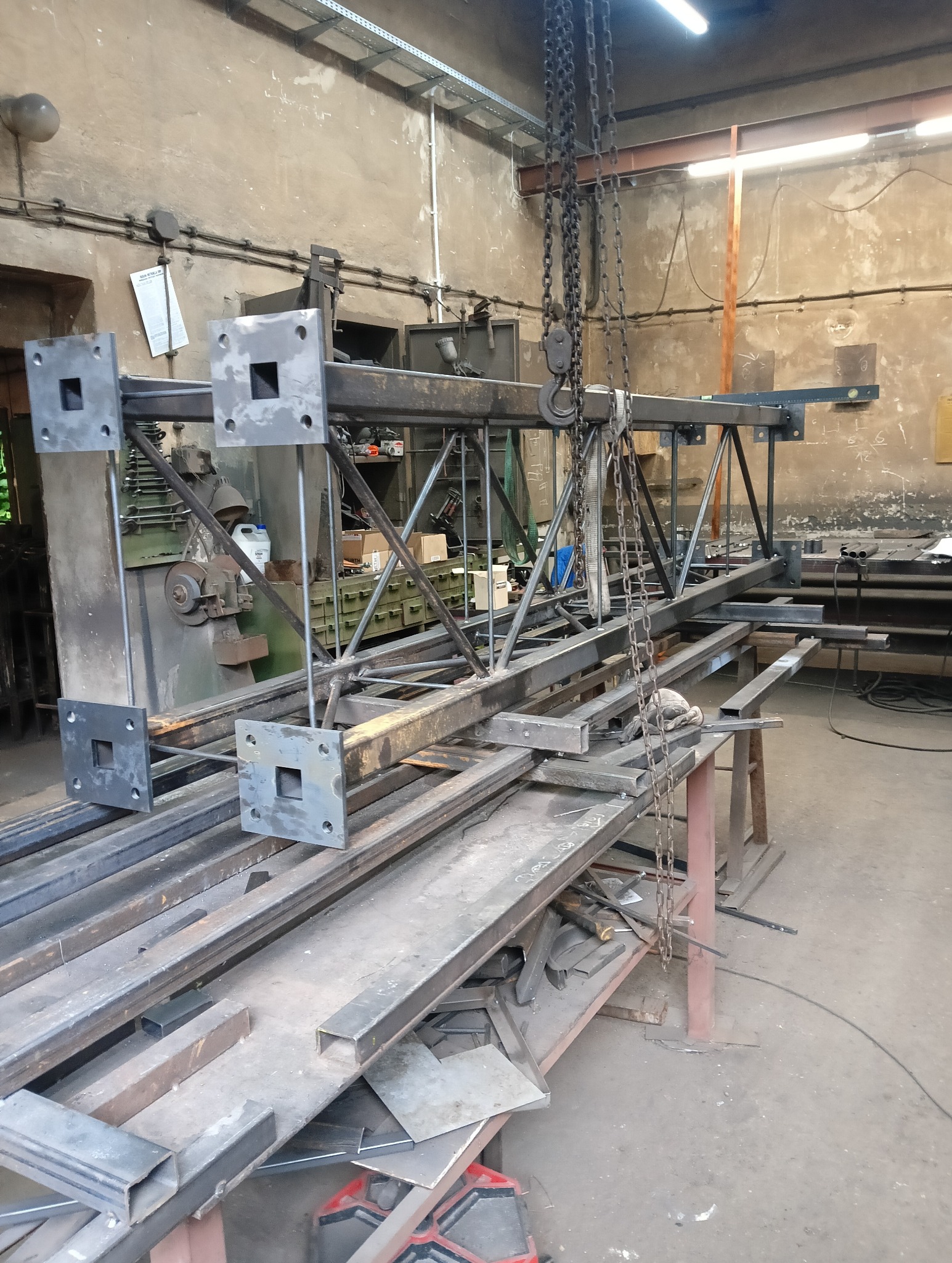 Konstrukcja stalowa z kwadratowymi elementami, podwieszona na łańcuchu w warsztacie. Widoczne profile i elementy spawane. Surowe wykończenie, warsztatowe otoczenie.