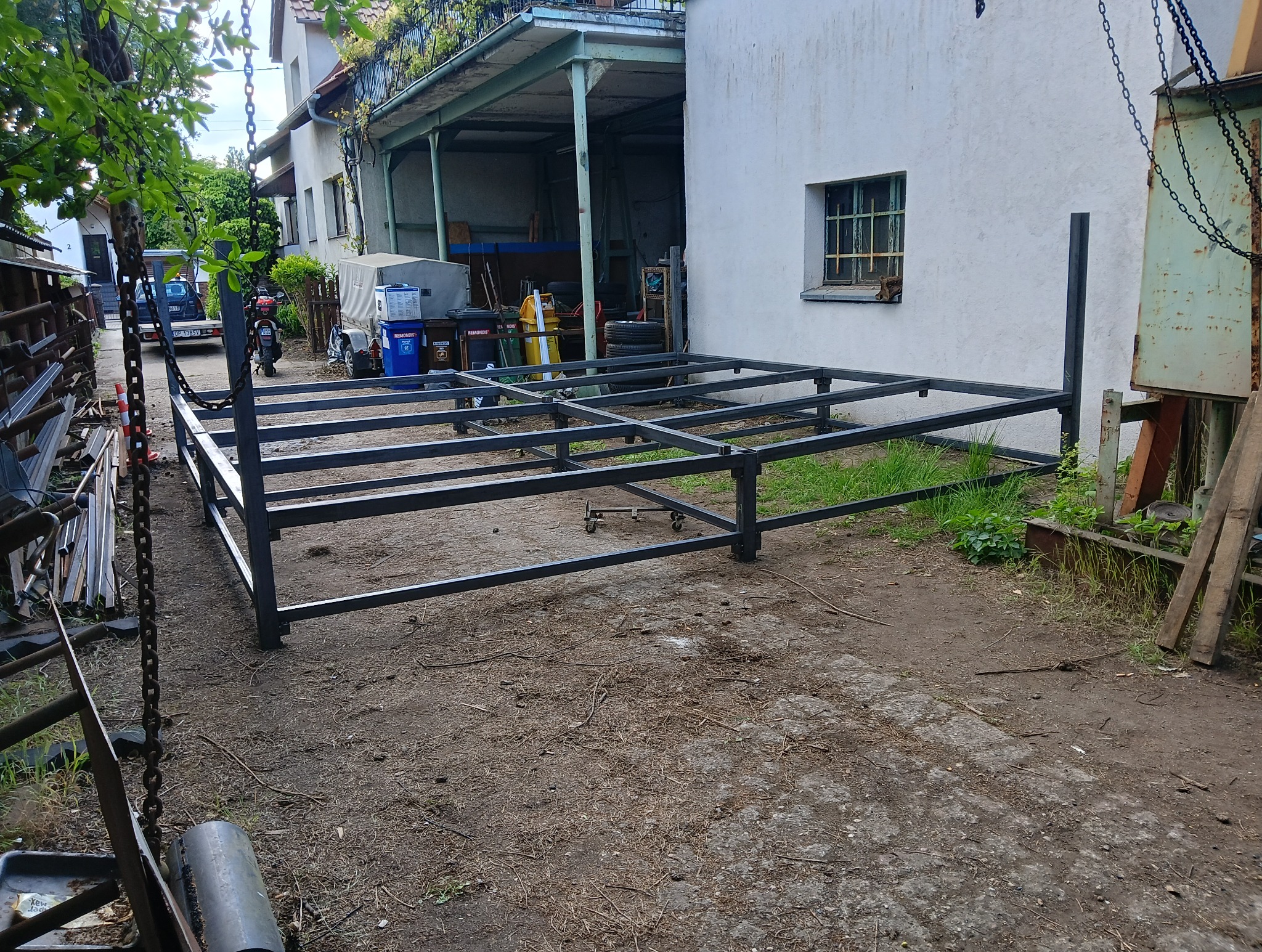 Konstrukcja metalowa, prawdopodobnie pod pergolę, w trakcie budowy. Widoczny szkielet z profili, tło z budynkami i zielenią.