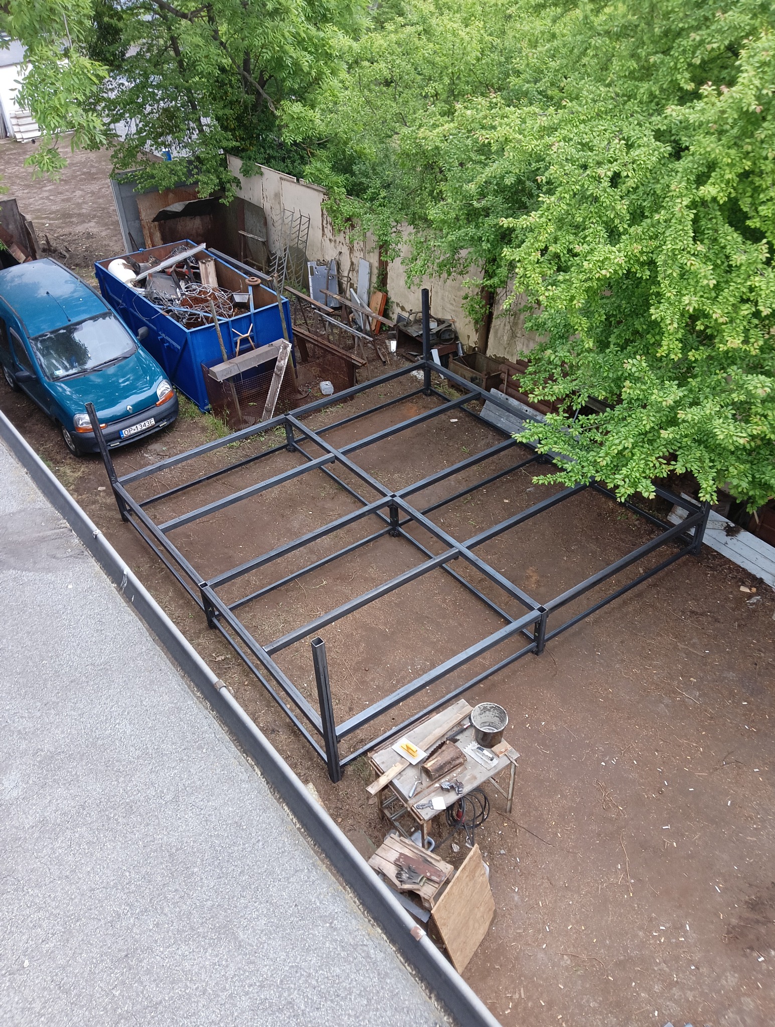 Metalowa konstrukcja szkieletowa w ogrodzie, widok z góry. Na ziemi leżą narzędzia. W tle zaparkowany samochód i kontener ze złomem.