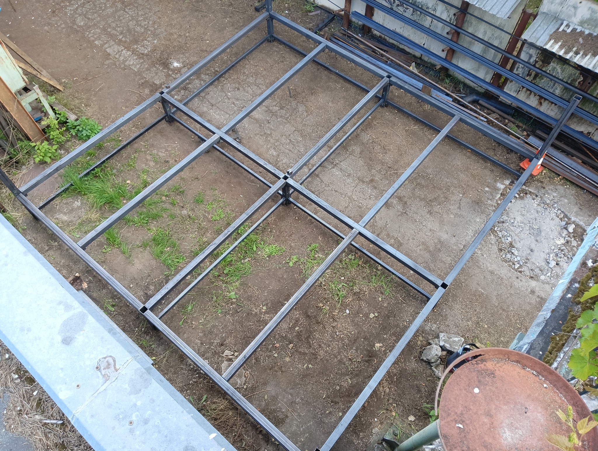 Metalowa konstrukcja szkieletowa z profili, prawdopodobnie pod zabudowę, widziana z góry, na ziemi z fragmentami trawy i betonu.