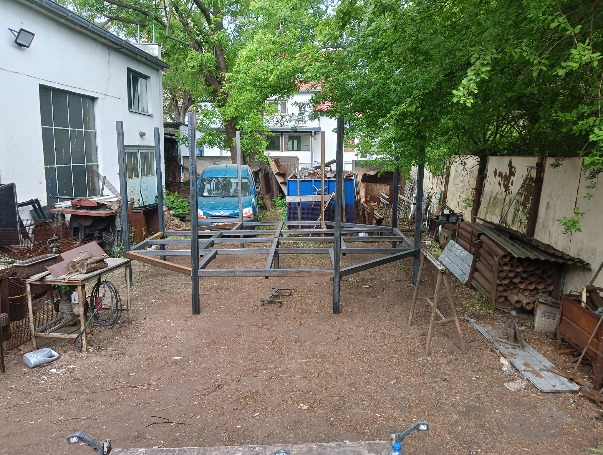 Stalowa konstrukcja szkieletowa w trakcie budowy na zewnątrz, z widocznymi elementami warsztatu i zielenią w tle. Konstrukcja spawana, surowy metal.