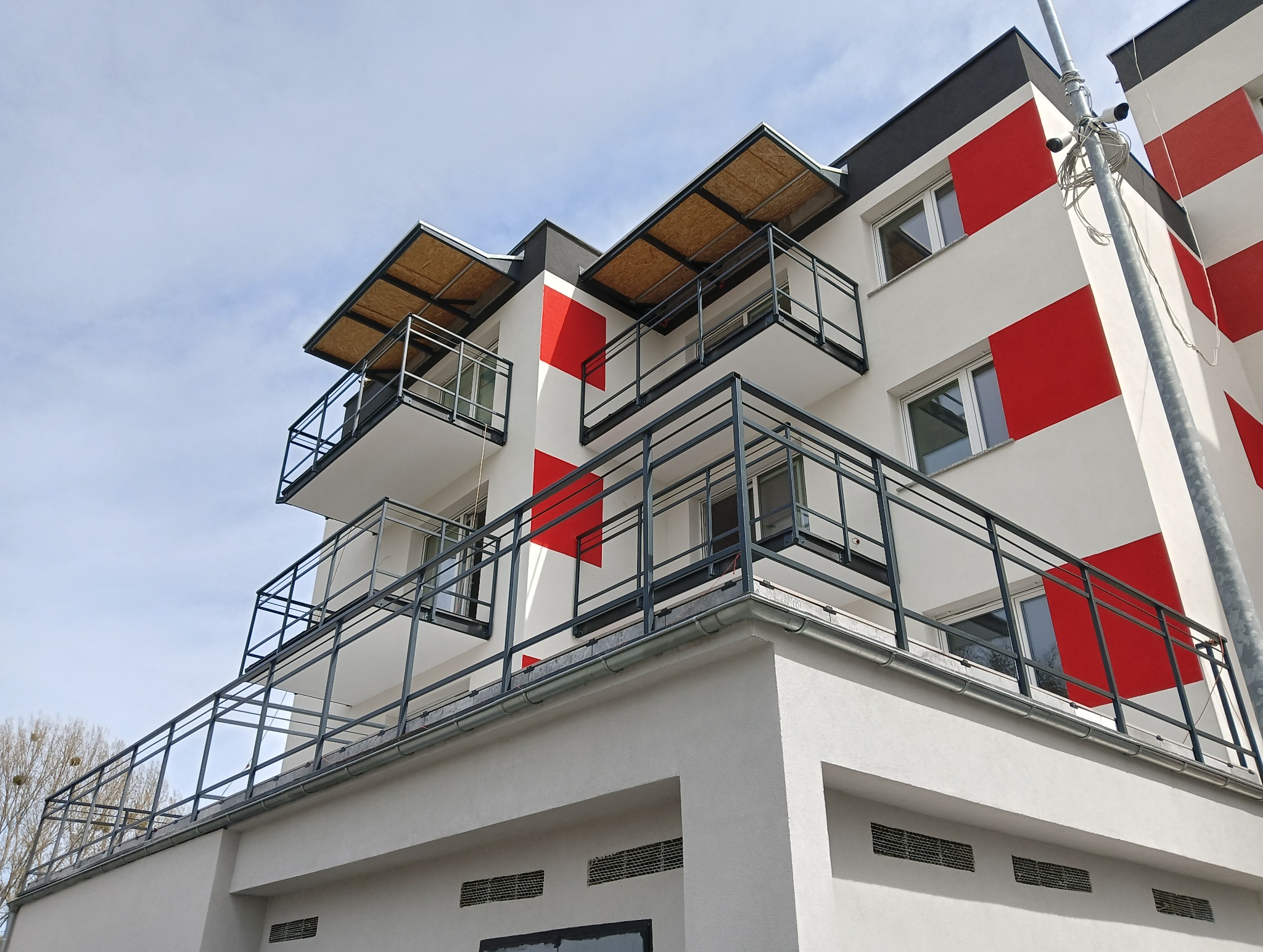 Nowoczesny budynek z balkonami z metalowymi balustradami w geometryczne wzory czerwieni i bieli. Widok z dołu w słoneczny dzień, podkreślający minimalistyczny design.