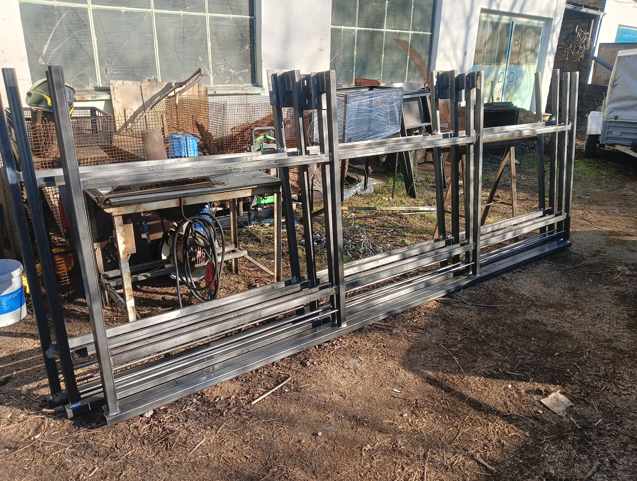 Stalowe ramy konstrukcyjne z profili kwadratowych, ułożone warstwami na ziemi, w tle warsztat z narzędziami i maszynami, prawdopodobnie gotowe elementy do montażu ogrodzenia.