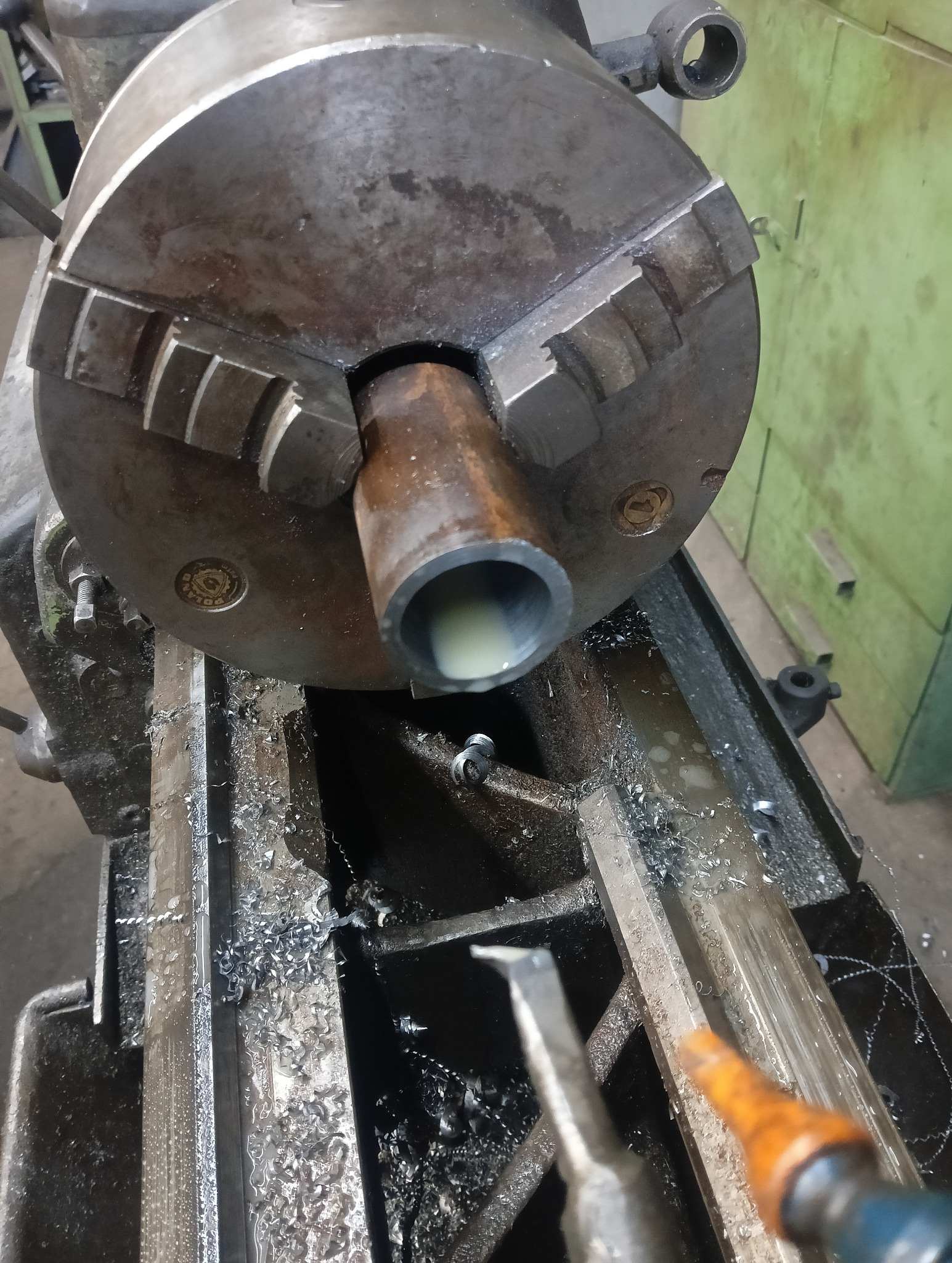 Metalowa rura w uchwycie tokarskim podczas obróbki, widoczne wióry i narzędzie skrawające. Szare kolory maszyny, chłodziwo wewnątrz rury, zbliżenie na proces.