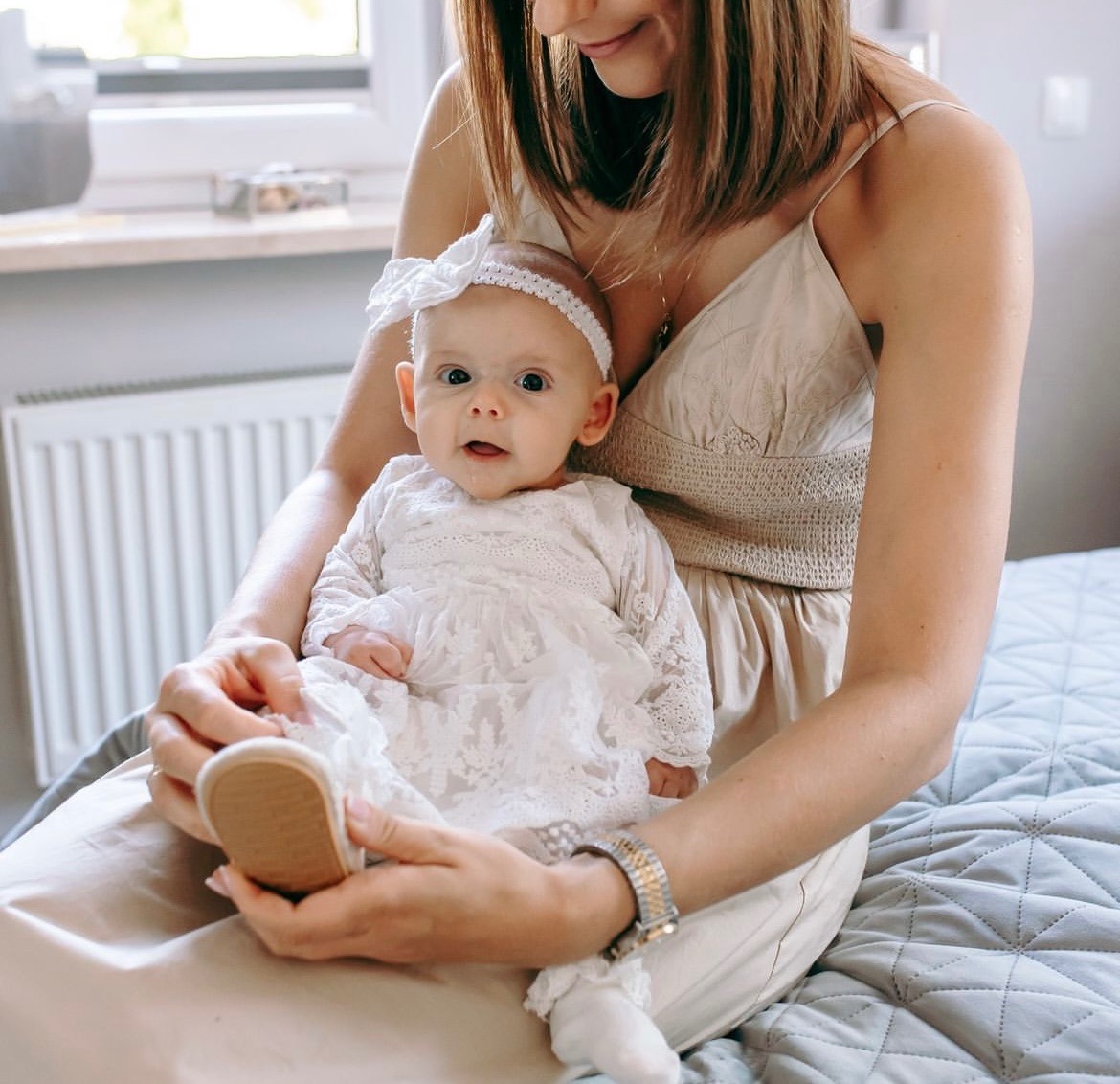 Matka trzymająca na kolanach niemowlę ubrane w białą, koronkową sukienkę i opaskę z kokardą, zakładająca mu beżowy bucik.
