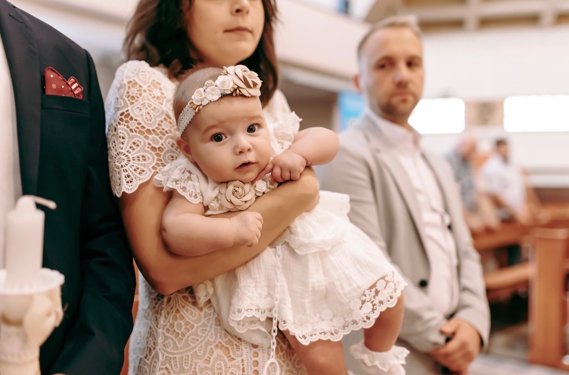Urocza dziewczynka w białej, koronkowej sukience i opasce z kwiatami, trzymana na rękach przez kobietę w kościele, w tle widoczny mężczyzna w jasnej marynarce.