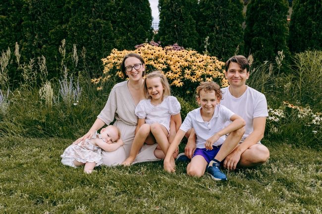 Uśmiechnięta rodzina z trójką dzieci pozuje na trawie przed kwitnącymi rabatami i tujami w tle.