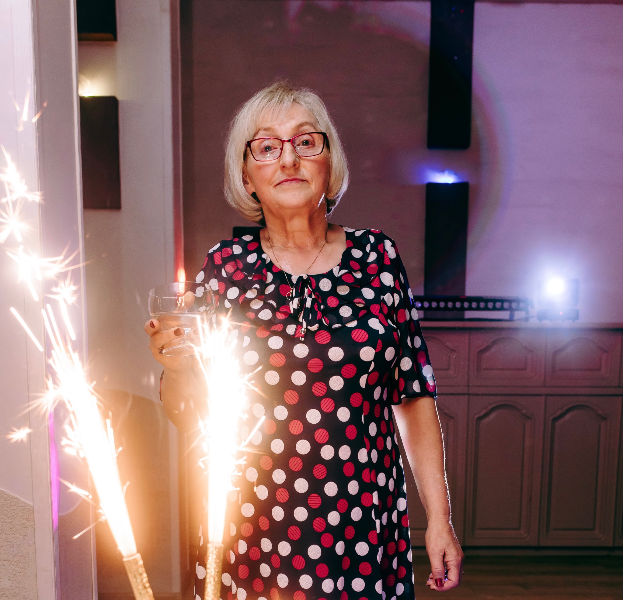 Uśmiechnięta kobieta w okularach i sukience w grochy trzyma kieliszek i zimne ognie w pomieszczeniu z oświetleniem eventowym.