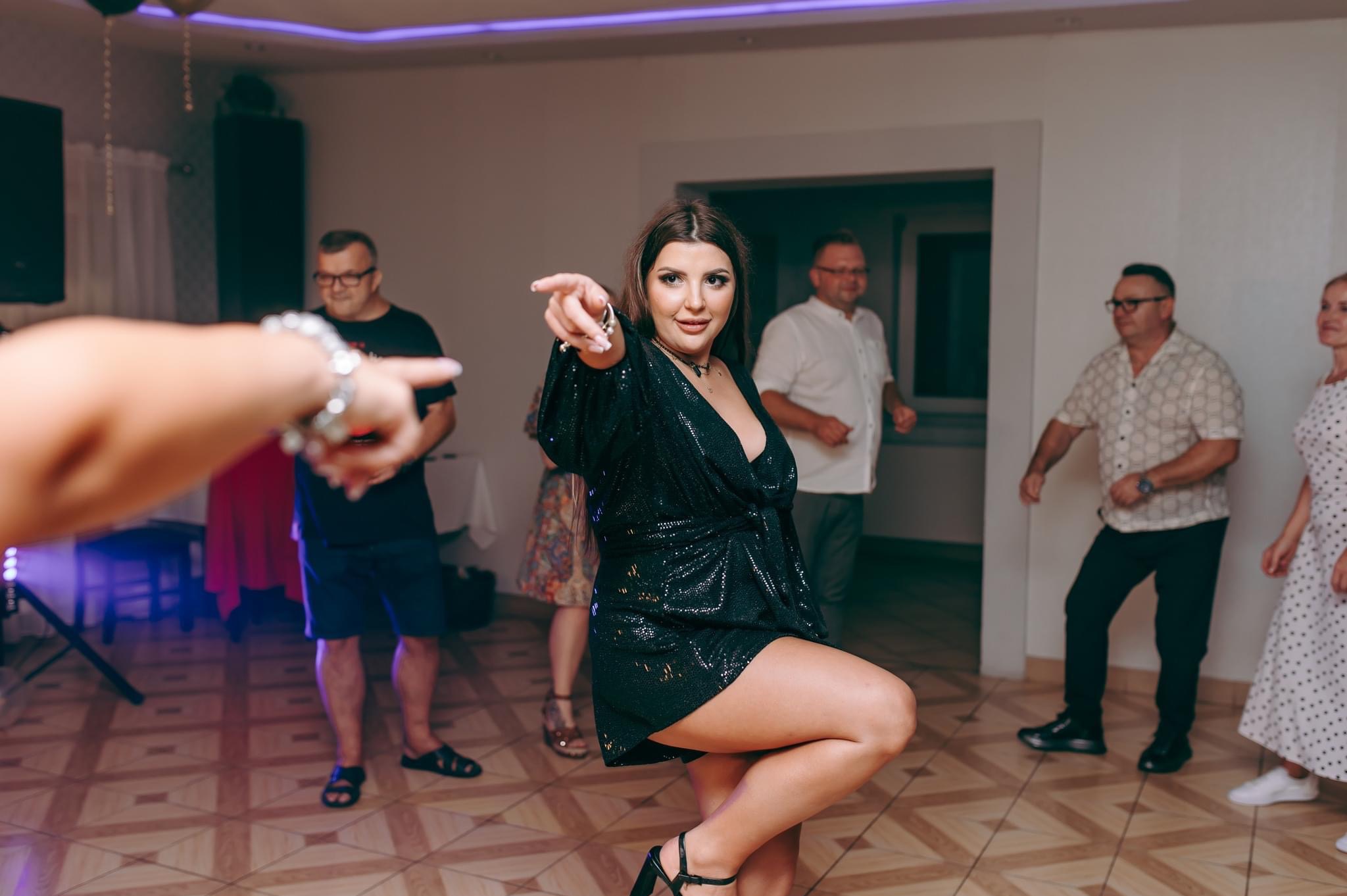 Kobieta w cekinowej sukience tańczy i wskazuje palcem w obiektyw podczas imprezy z innymi osobami w tle.