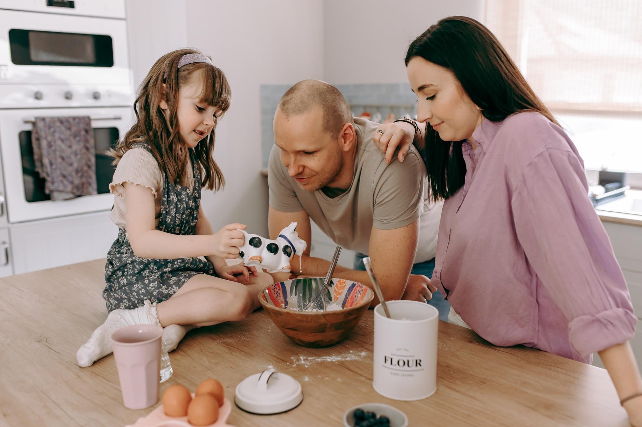 Rodzina w kuchni: córka w sukience w kwiaty wlewa mleko z dzbanka w kształcie krowy do miski, ojciec i matka obserwują, na blacie jajka, mąka i borówki.