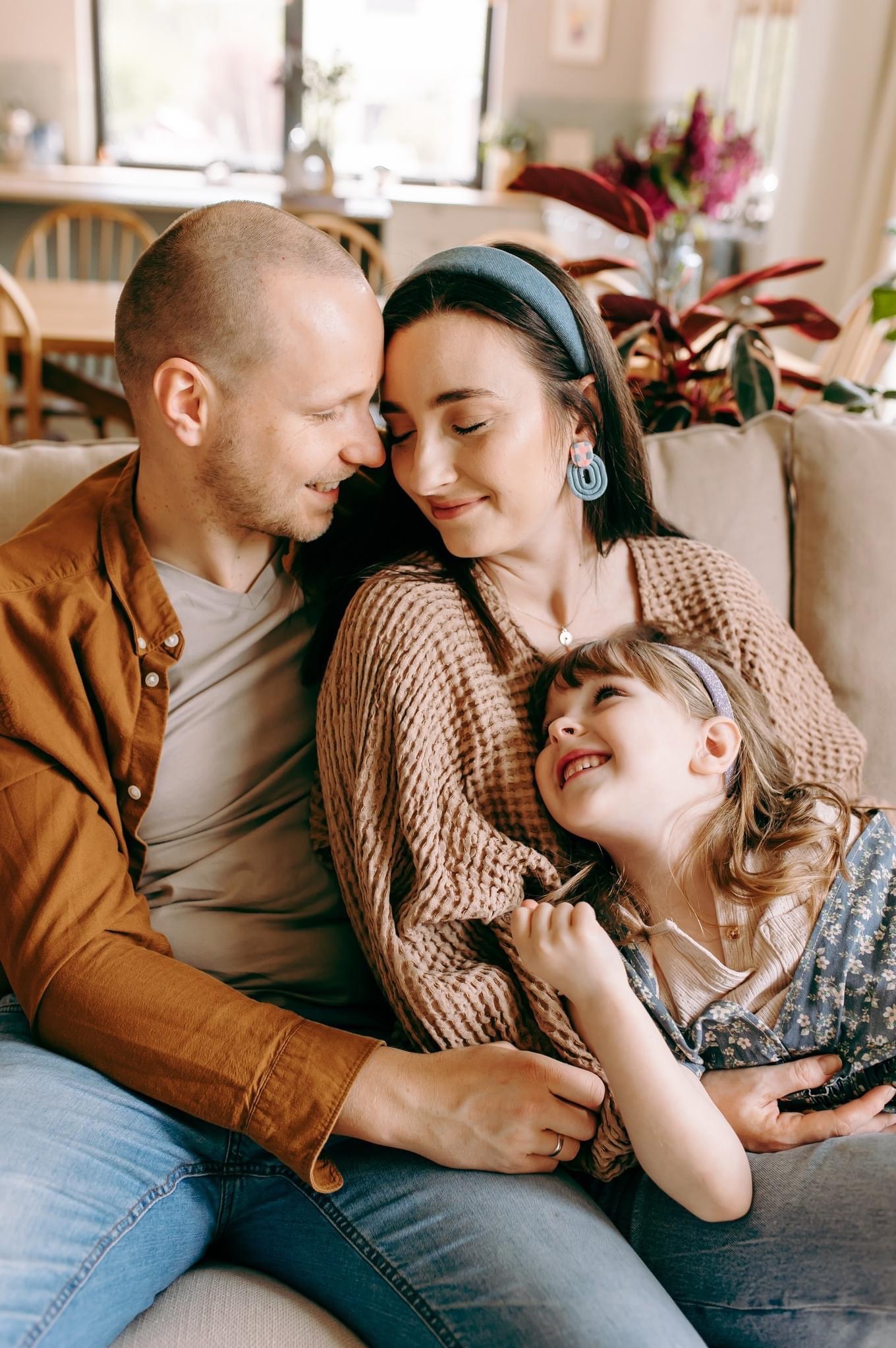 Szczęśliwa rodzina: tata, mama i córka siedzą blisko siebie na beżowej kanapie, patrząc na siebie z uśmiechem; kobieta ma na głowie niebieską opaskę i kolczyki w kształcie geometrycznym,...