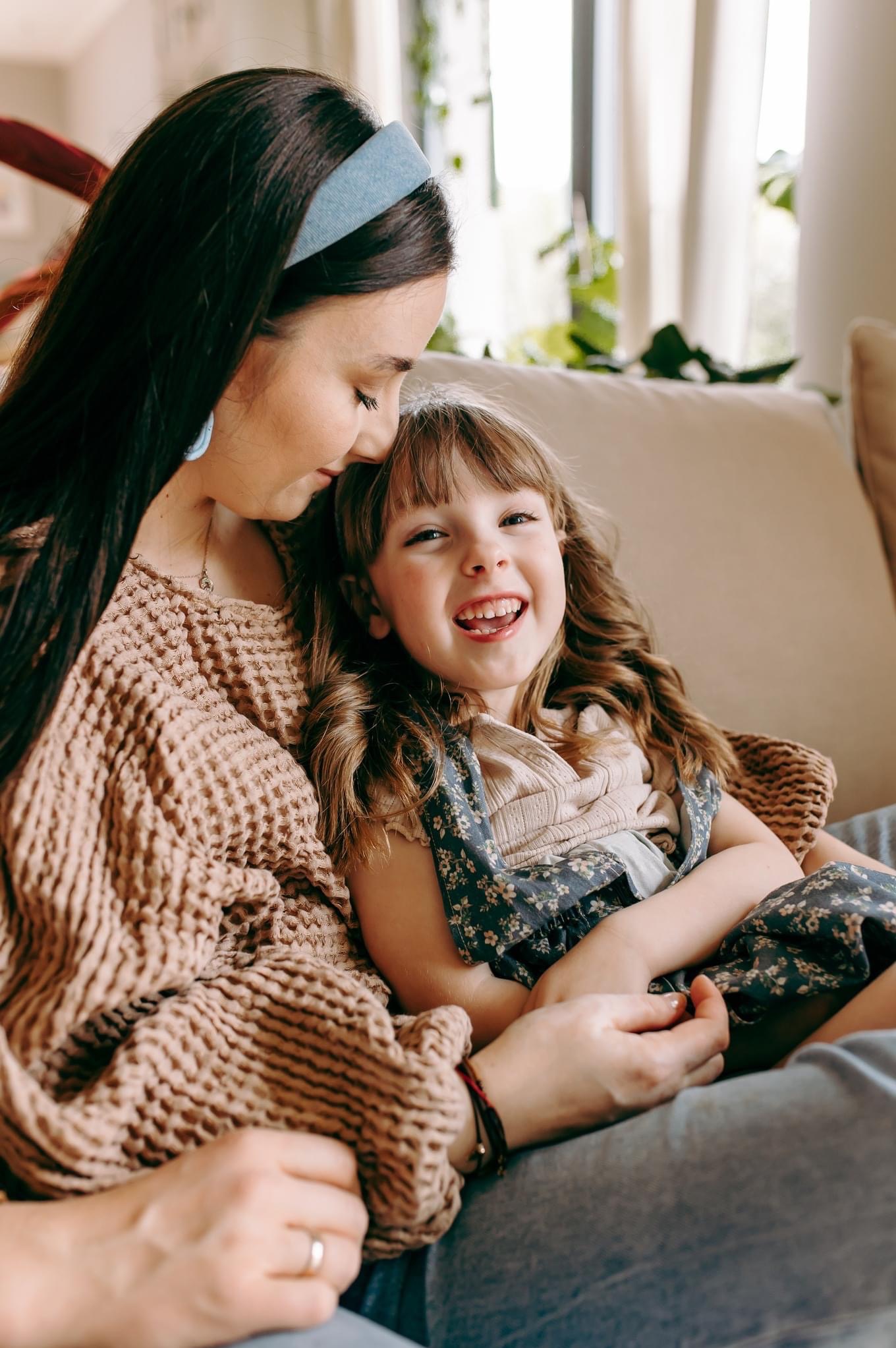 Uśmiechnięta dziewczynka w kwiecistej sukience siedzi na kolanach mamy, która ma na głowie błękitną opaskę i sweter o grubym splocie. Matka patrzy z czułością na córkę.
