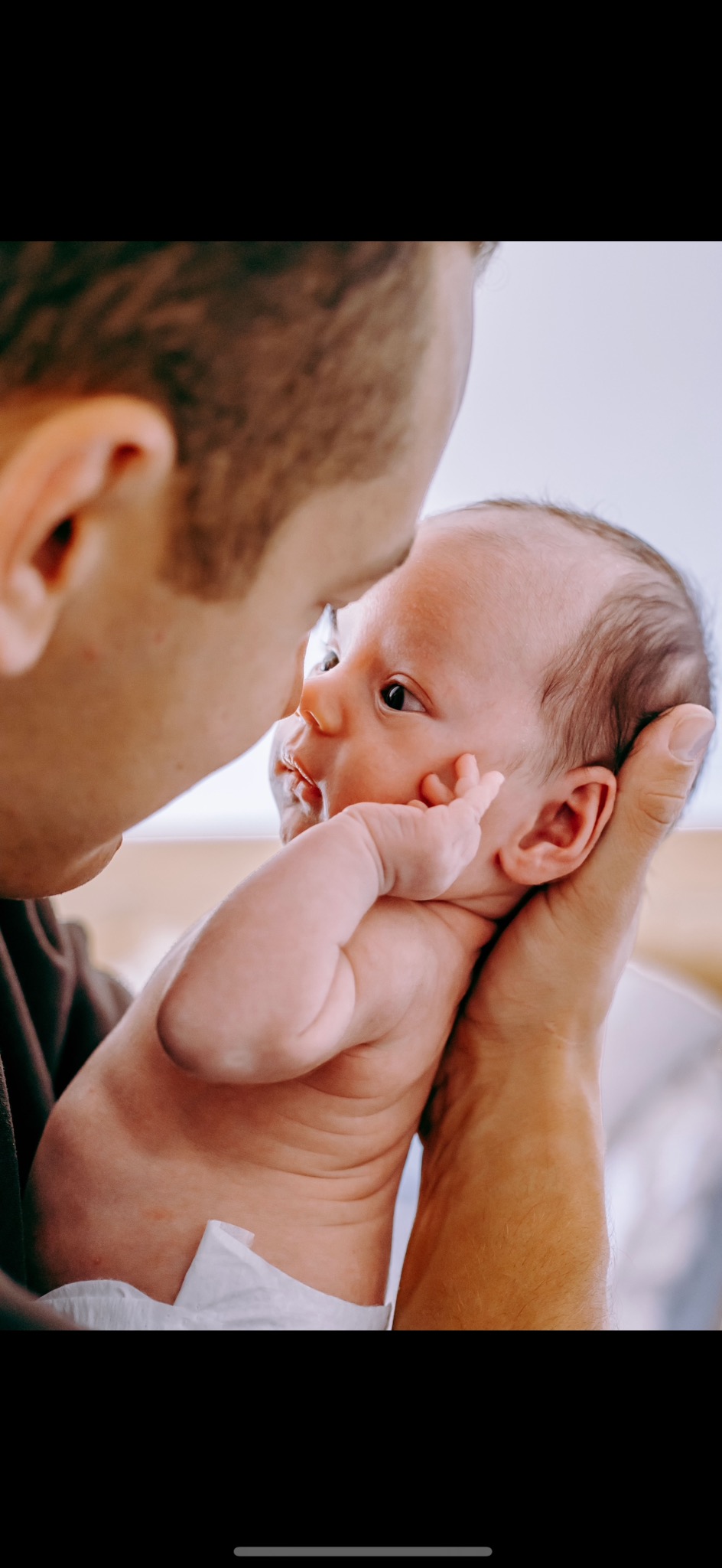 Ojciec trzyma noworodka, który patrzy z zaciekawieniem, z małą rączką przy twarzy, w miękkim, naturalnym świetle.