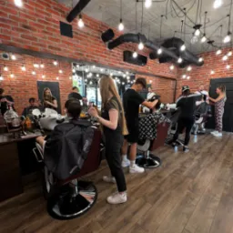 Wnętrze lokalu z ceglanymi ścianami i licznymi żarówkami, gdzie kilku pracowników obsługuje klientów na fotelach fryzjerskich, wykonując strzyżenie i inne zabiegi.