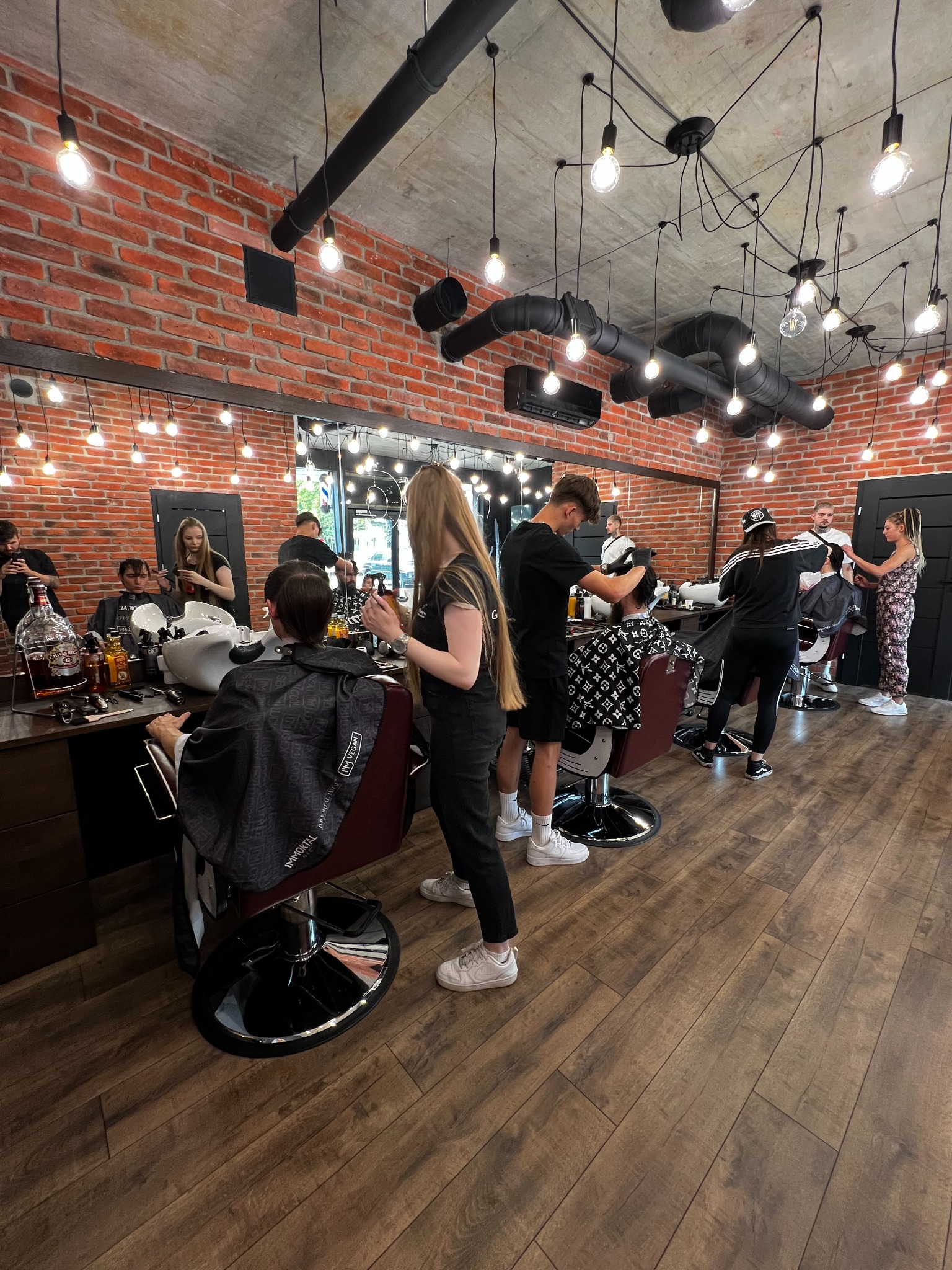 Wnętrze lokalu z ceglanymi ścianami i licznymi żarówkami, gdzie kilku pracowników obsługuje klientów na fotelach fryzjerskich, wykonując strzyżenie i inne zabiegi.