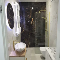 Elegancka łazienka z marmurową ścianą, złotą armaturą, owalnym lustrem z podświetleniem LED i białym grzejnikiem drabinkowym.