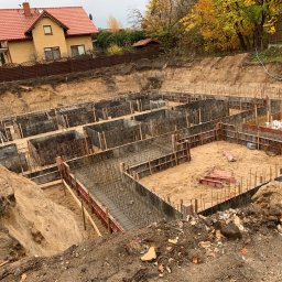 MAK-BUD DEVELOPMENT - Zalane fundamenty domu jednorodzinnego z widoczną stalą zbrojeniową i szalunkami, na tle wykopu i budynku w jesiennej scenerii. Ujęcie z góry.
