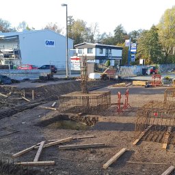 MAK-BUD DEVELOPMENT - Zbrojenie fundamentów pod budowę, widoczne stalowe konstrukcje w wykopach, przygotowane do zalania betonem. W tle budynki i drzewa.