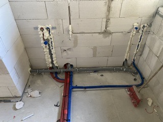 Instalacja hydrauliczna w surowym stanie: rury PEX (czerwone i niebieskie) prowadzone wzdłuż ściany z betonu komórkowego, zabezpieczone pianką montażową, widoczne przyłącza i elementy mocujące.