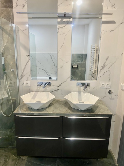 Nowoczesna łazienka z dwiema geometrycznymi umywalkami nablatowymi, chromowanymi bateriami i lustrami na marmurowej ścianie.