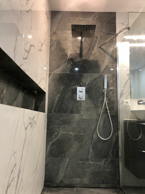 Nowoczesna kabina prysznicowa z deszczownicą i baterią podtynkową, wykończona płytkami imitującymi marmur w odcieniach szarości i bieli, z widoczną wnęką w ścianie.