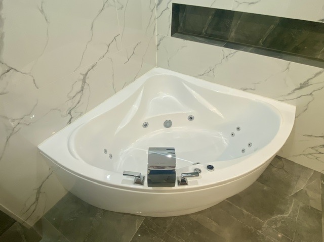 Biała narożna wanna z hydromasażem i chromowaną armaturą, otoczona marmurowymi płytkami ściennymi i podłogowymi w odcieniach szarości.