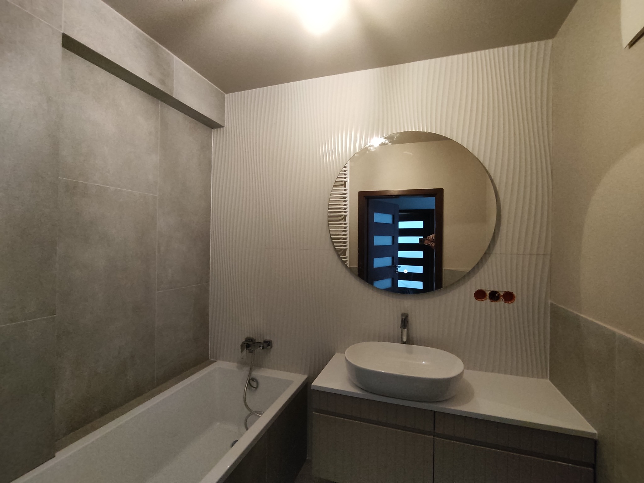 Nowoczesna łazienka z wanną, umywalką nablatową i okrągłym lustrem, ściany wykończone płytkami w odcieniach szarości i bieli z delikatnym wzorem fal, widoczne wystające kable elektryczne.