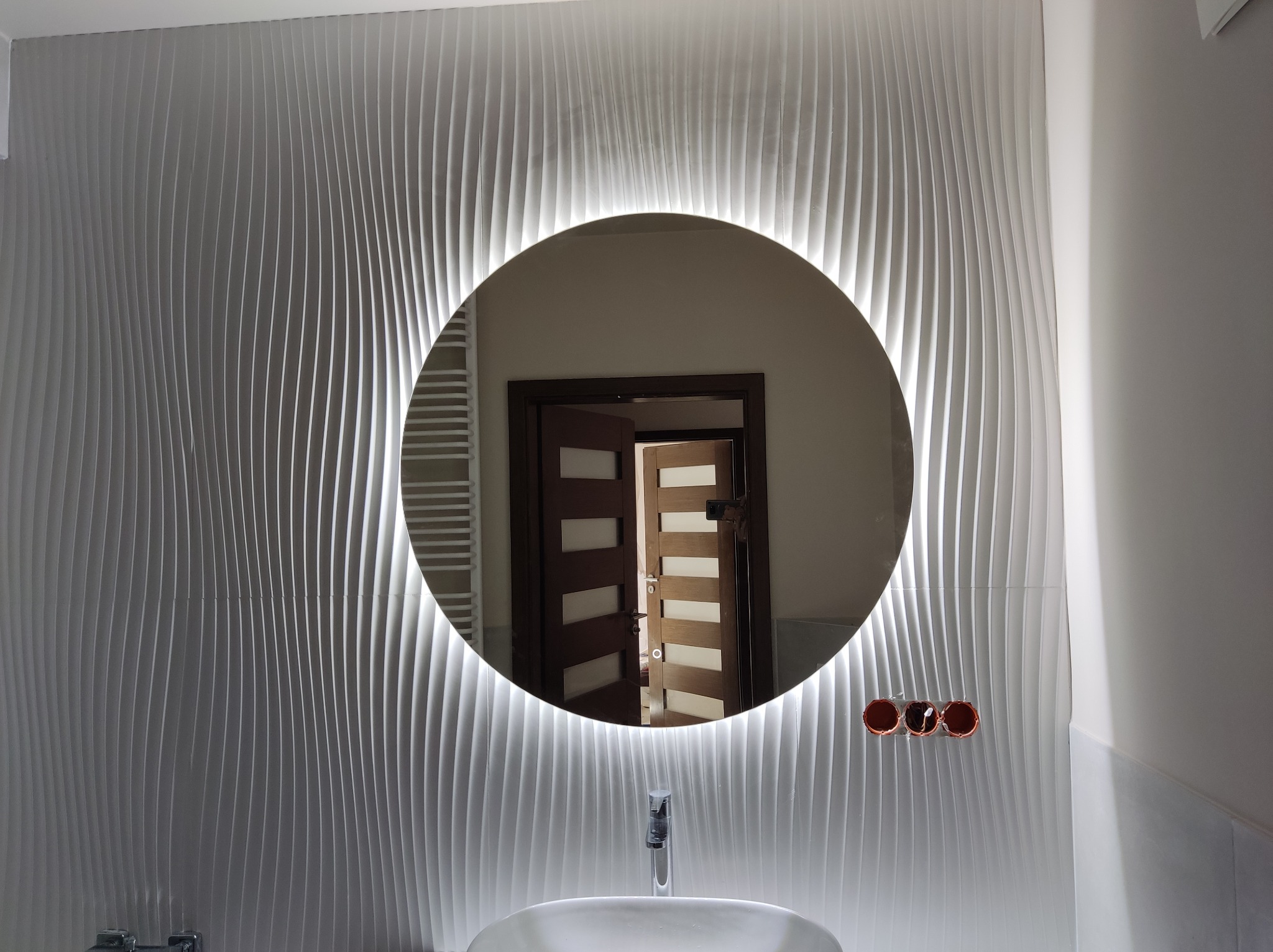 Nowoczesna łazienka z umywalką, okrągłym lustrem z podświetleniem LED i ścianą wykończoną trójwymiarowymi panelami dekoracyjnymi; w lustrze widoczne odbicie otwartych drzwi.