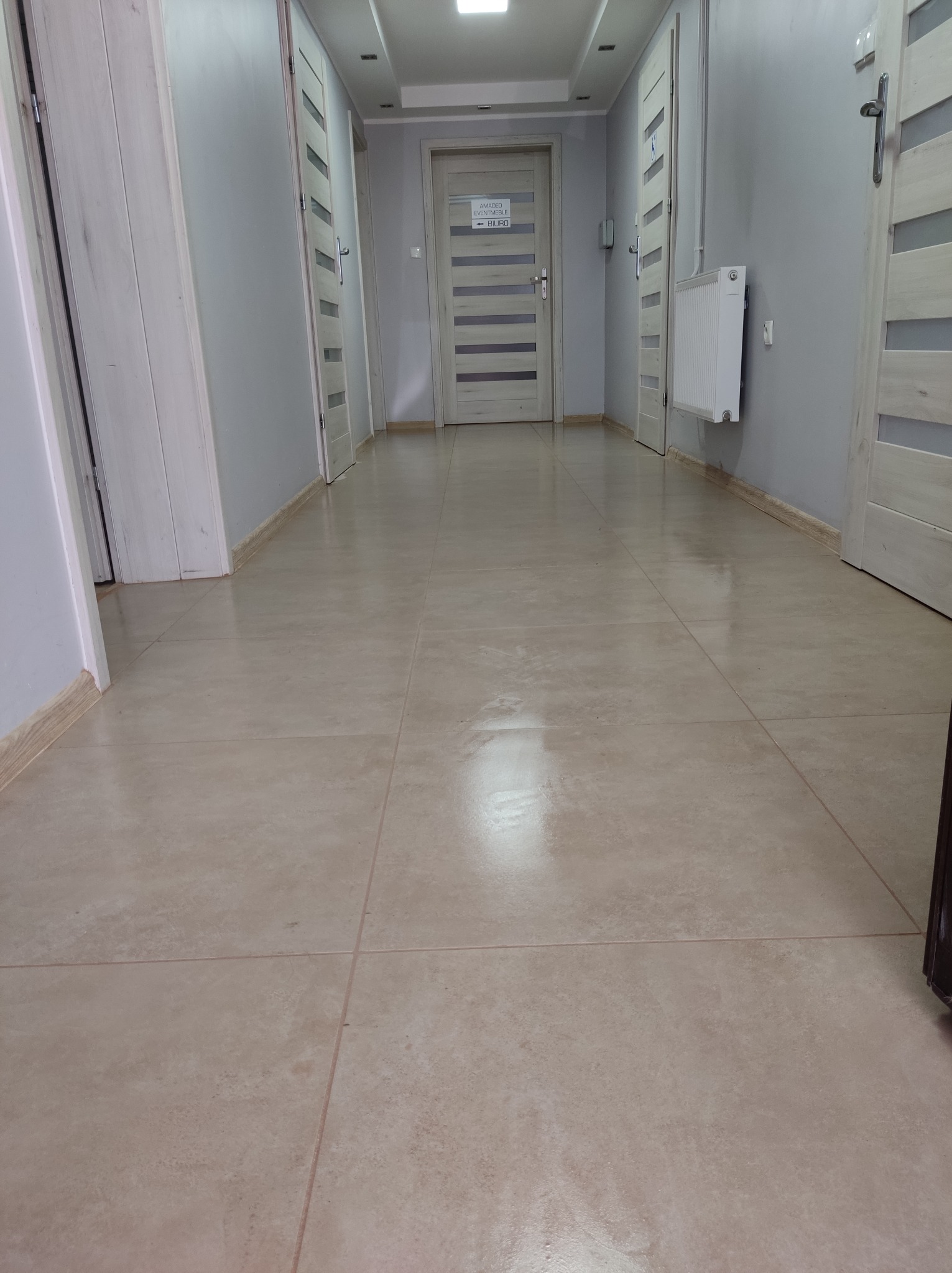 Korytarz z beżową podłogą z płytek, szare ściany i białe drzwi z poziomymi przeszkleniami, widoczny grzejnik na ścianie.