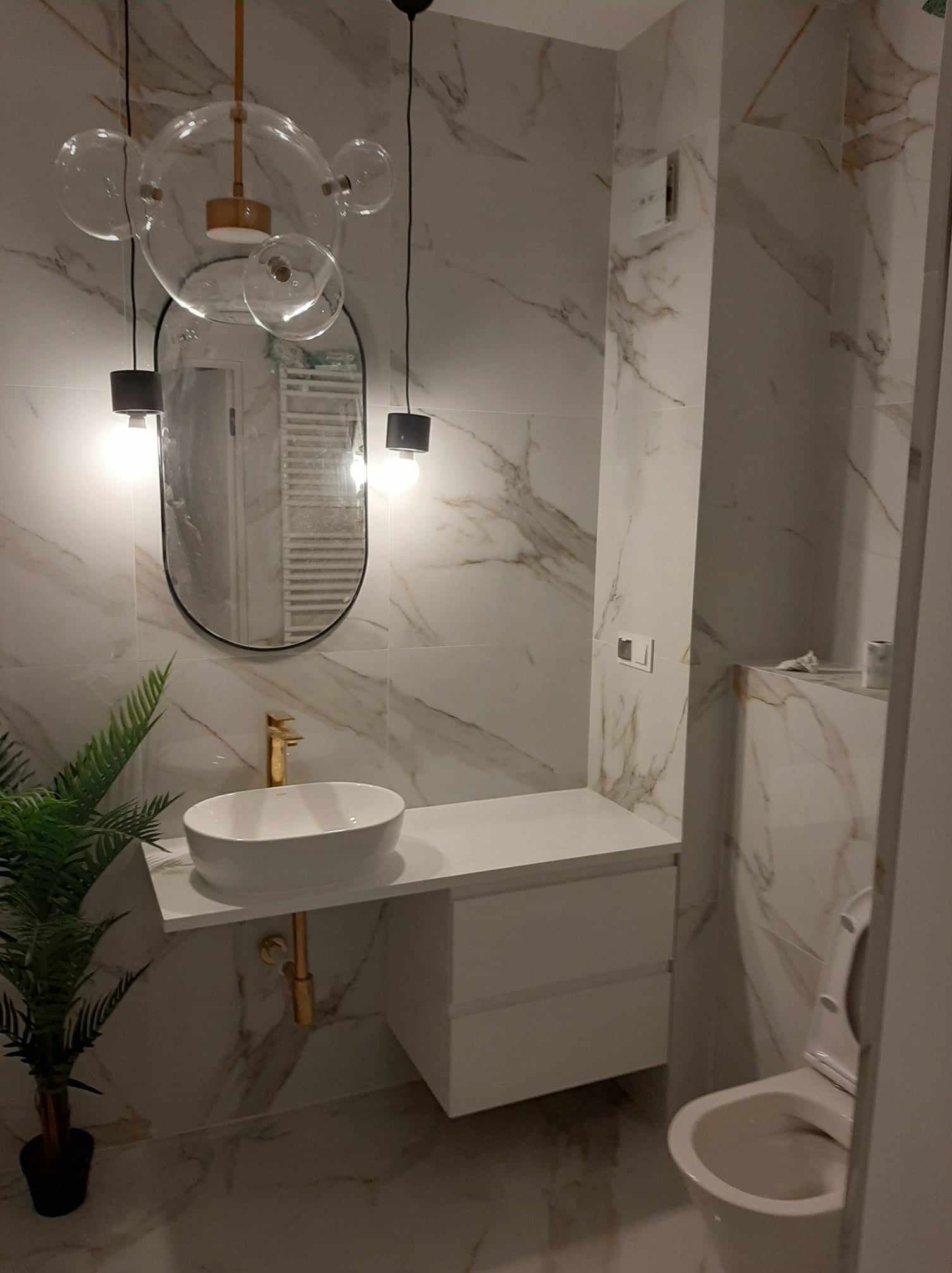 Nowoczesna łazienka w Wygrance z marmurowymi płytkami, owalną umywalką na białym blacie, złotą armaturą i designerską lampą wiszącą z szklanych kul, odbijającą się w owalnym lustrze.