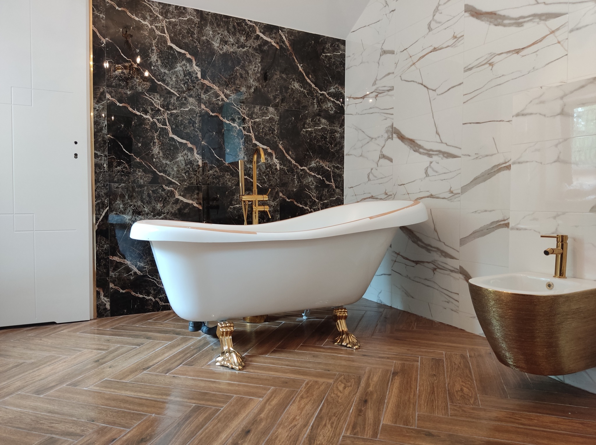 Elegancka łazienka z wolnostojącą białą wanną na złotych nóżkach, złotą armaturą i bidetem, wykończona płytkami imitującymi marmur w kolorach czerni i bieli, oraz drewnopodobną podłogą ułożoną...