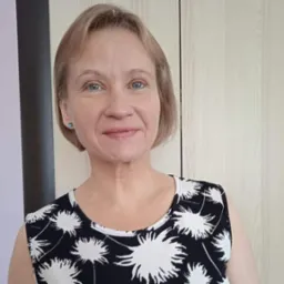 Portret uśmiechniętej kobiety o krótkich blond włosach i niebieskich oczach, ubrana w czarno-białą sukienkę w kwiaty, stojąca na tle jasnych drzwi.