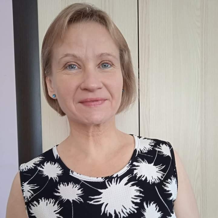 Portret uśmiechniętej kobiety o krótkich blond włosach i niebieskich oczach, ubrana w czarno-białą sukienkę w kwiaty, stojąca na tle jasnych drzwi.