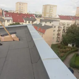 Widok z góry na płaski dach pokryty papą, z nowo zamontowaną, metalową obróbką blacharską na krawędzi. Na dachu leżą rolki papy w kartonie, a w tle widać zabudowę miejską.