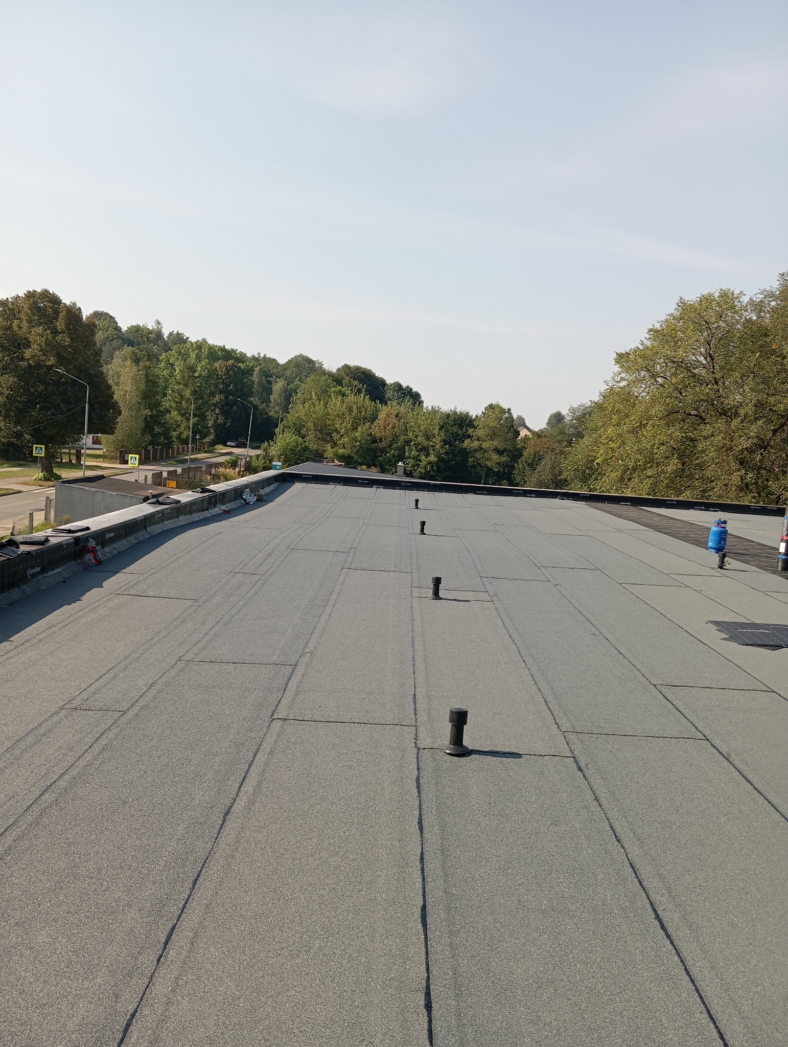 Wykończony, płaski dach pokryty papą termozgrzewalną, z widocznymi kominkami wentylacyjnymi i butlą gazową w tle, otoczony zielenią drzew.