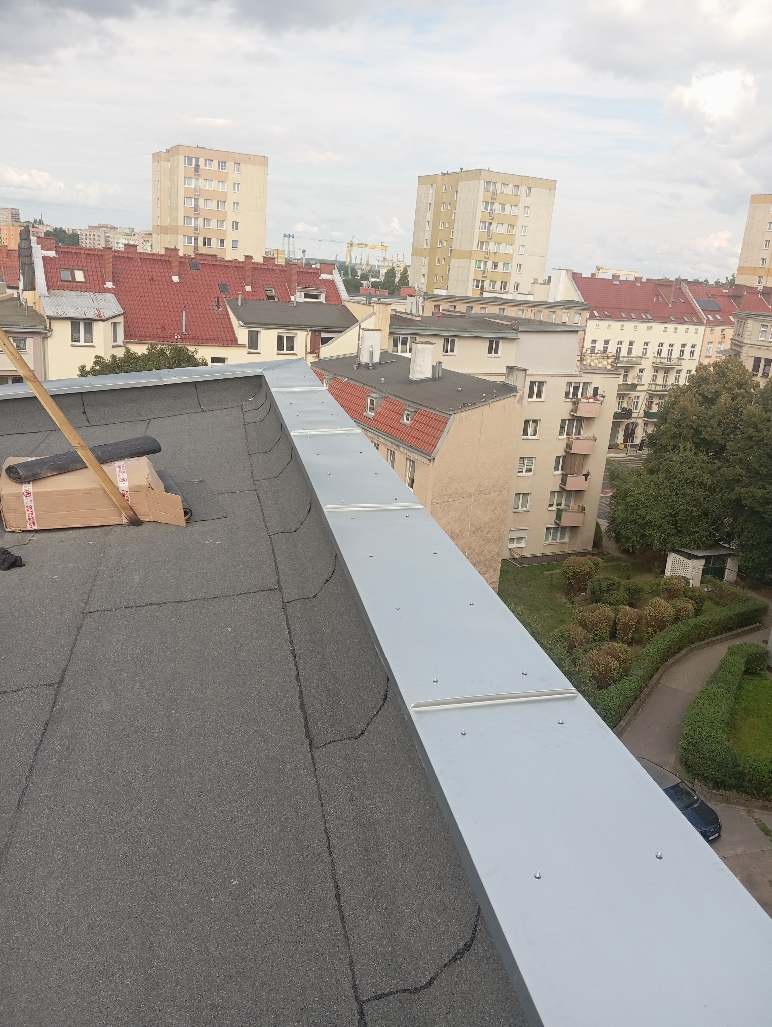 Widok z góry na płaski dach pokryty papą, z nowo zamontowaną, metalową obróbką blacharską na krawędzi. Na dachu leżą rolki papy w kartonie, a w tle widać zabudowę miejską.