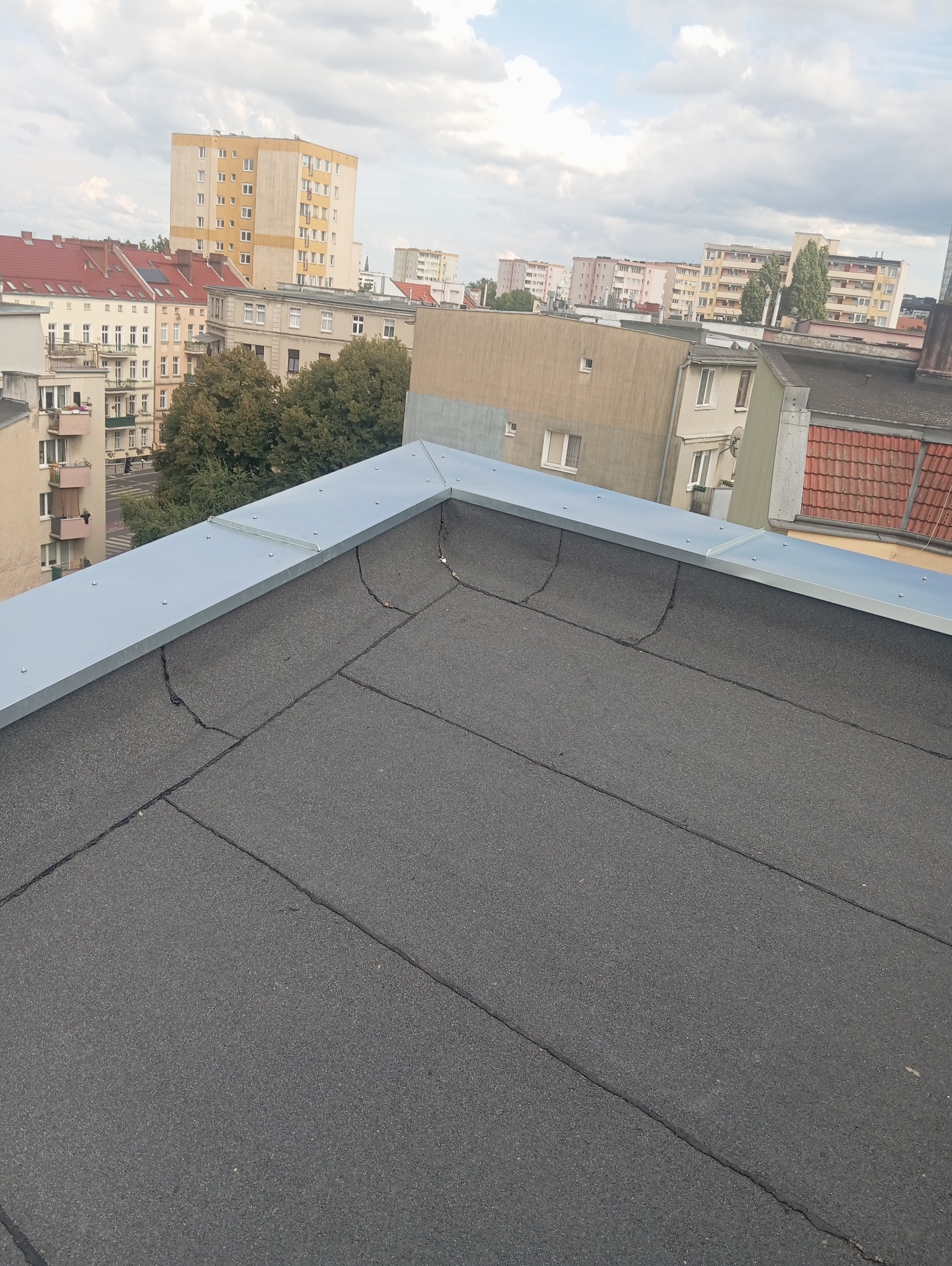 Płaski dach pokryty papą z widocznymi pęknięciami, obróbka blacharska w kolorze szarym, w tle zabudowa miejska i pochmurne niebo.