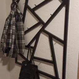 Czarny, geometryczny wieszak metalowy przymocowany do ściany, z zawieszoną koszulą w kratę i czarną torbą.