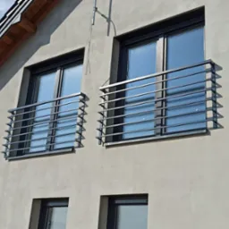 Dwa okna z metalowymi, poziomymi balustradami na elewacji domu. Balustrady w kolorze grafitowym, tło stanowi jasna elewacja i błękitne niebo.