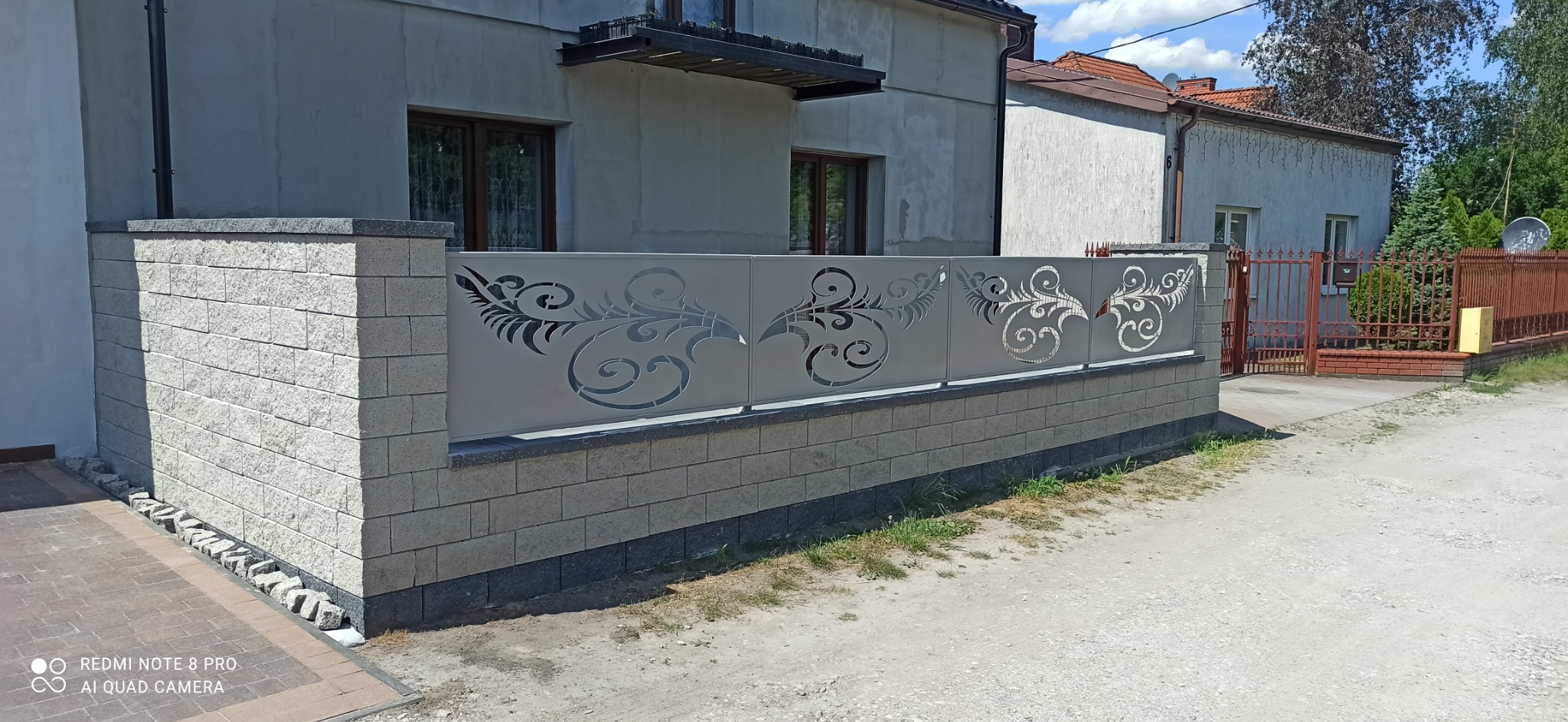 Nowoczesne ogrodzenie panelowe z dekoracyjnym ażurowym wzorem roślinnym, zamontowane na niskim murku z szarej cegły.