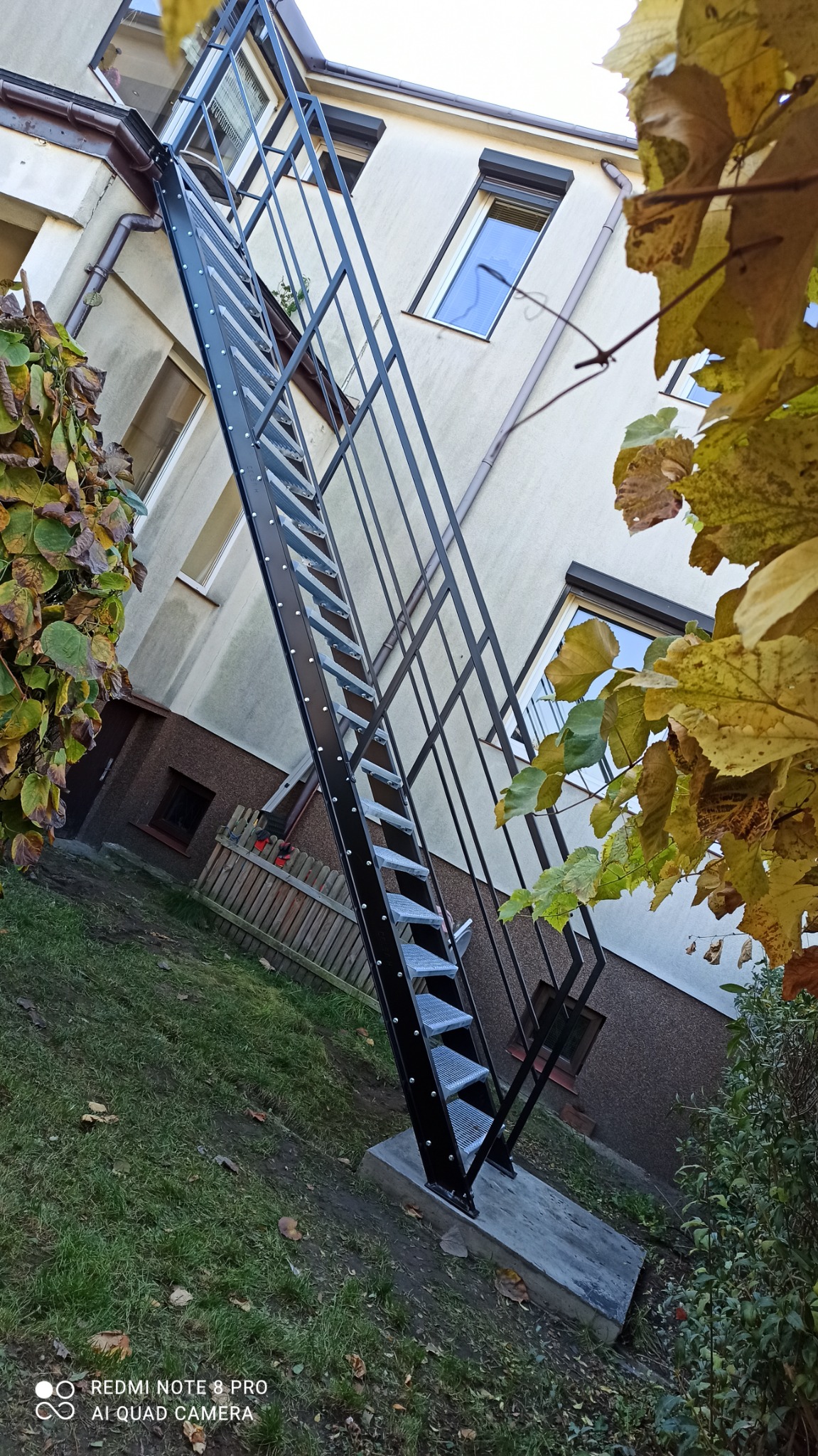 Zewnętrzne, metalowe schody ewakuacyjne przy budynku mieszkalnym, widok z dołu, z liśćmi drzew na pierwszym planie.