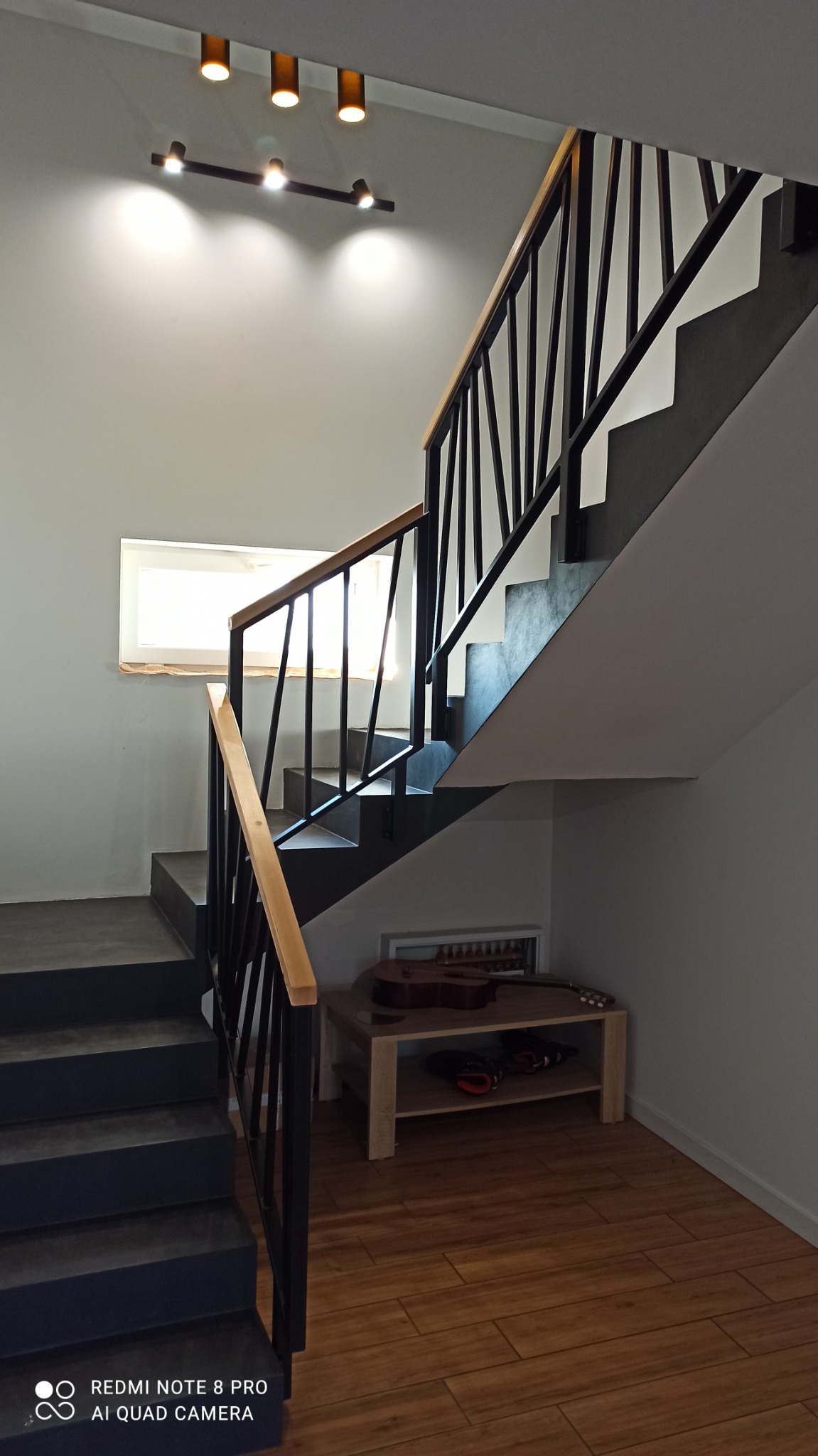 Nowoczesne schody z czarnymi metalowymi poręczami i drewnianym uchwytem, oświetlone punktowymi lampami sufitowymi, z widocznym fragmentem okna i drewnianą podłogą, pod schodami stolik z gitarą.