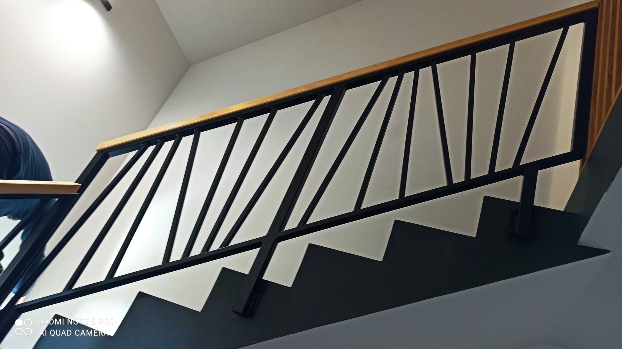 Nowoczesne schody metalowe z czarną konstrukcją i drewnianą poręczą, widok z dołu, z fragmentem osoby na pierwszym planie.